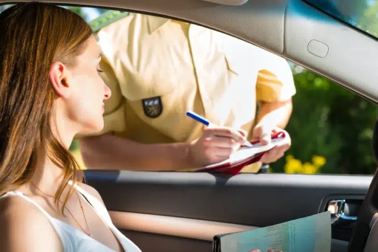 A woman in a car receiving a traffic ticket from a police officer writing on a clipboard outside the driver's window.