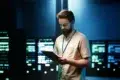 Man with beard in tan shirt holding tablet in front of glowing server racks.