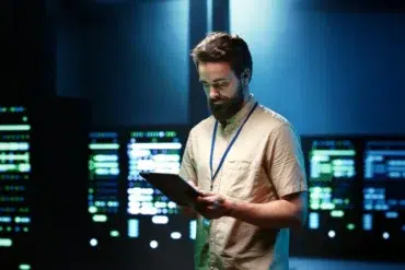Man with beard in tan shirt holding tablet in front of glowing server racks.