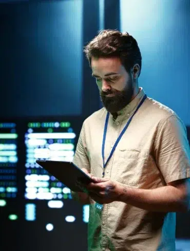 Man with beard in tan shirt holding tablet in front of glowing server racks.