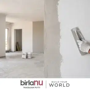 A gloved hand applies putty to a white wall in a spacious, unfinished room with large windows. Two bags of putty are on the floor. Calm, constructive atmosphere.