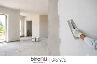 A gloved hand applies putty to a white wall in a spacious, unfinished room with large windows. Two bags of putty are on the floor. Calm, constructive atmosphere.