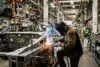 A welder in a mask and protective clothing sparks bright blue and orange while joining metal car frame parts on an assembly line.