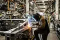 A welder in a mask and protective clothing sparks bright blue and orange while joining metal car frame parts on an assembly line.