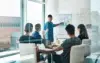 A male presenter points to a whiteboard with sticky notes and flowcharts while a diverse group of colleagues watches in a modern meeting room.
