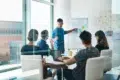 A male presenter points to a whiteboard with sticky notes and flowcharts while a diverse group of colleagues watches in a modern meeting room.