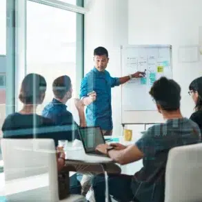 A male presenter points to a whiteboard with sticky notes and flowcharts while a diverse group of colleagues watches in a modern meeting room.