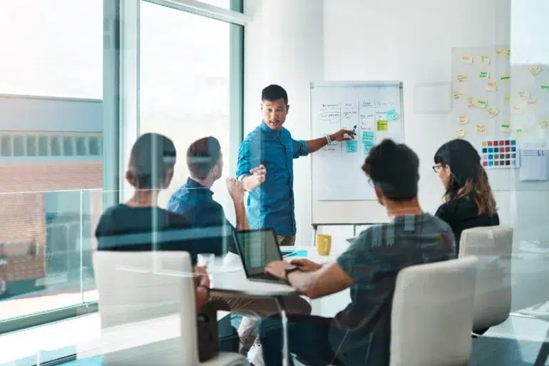 A male presenter points to a whiteboard with sticky notes and flowcharts while a diverse group of colleagues watches in a modern meeting room.