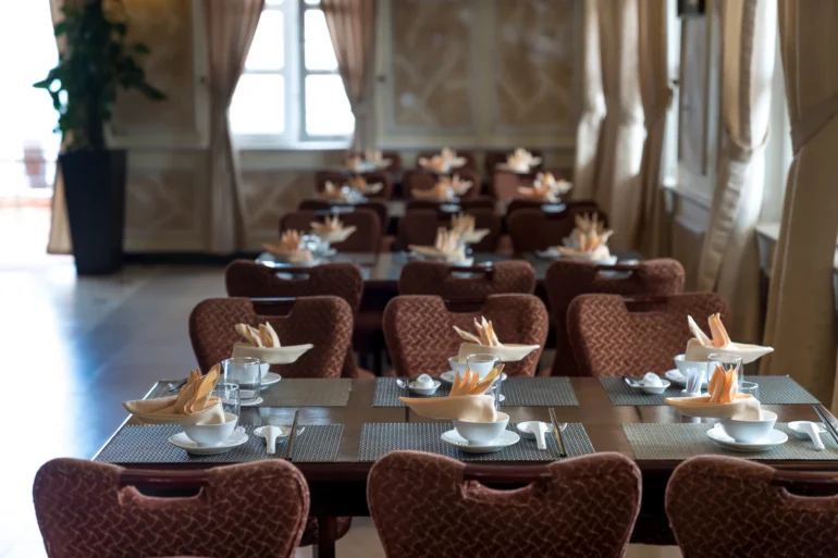 Rows of dark wooden dining tables with place settings featuring folded napkins in bowls and patterned chairs in a restaurant with large windows.