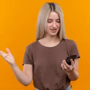 Young woman with blonde hair and braces looking at her phone and gesturing with her left hand against an orange background.