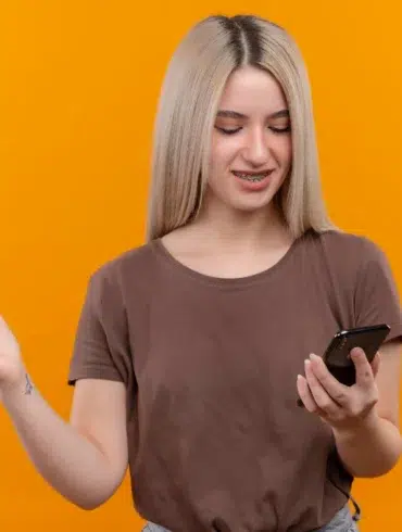 Young woman with blonde hair and braces looking at her phone and gesturing with her left hand against an orange background.