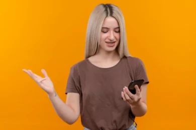 Young woman with blonde hair and braces looking at her phone and gesturing with her left hand against an orange background.