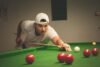 A young man wearing a white t-shirt and backwards cap leans over a green snooker table, lining up a shot with a cue stick among several red and white balls.