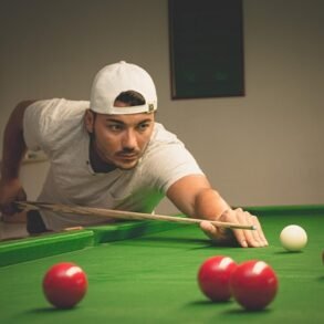 A young man wearing a white t-shirt and backwards cap leans over a green snooker table, lining up a shot with a cue stick among several red and white balls.