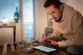 Man in beige hoodie writing in notebook at a wooden table with a tablet and glass of water.