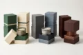 Three sets of stacked, textured rectangular boxes in green, cream, gray, and burgundy, with two open boxes displaying candles and one wooden vessel on a white background.