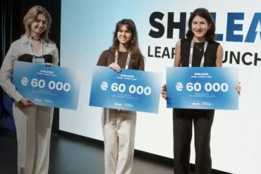 Three young women stand with signs announcing "SHELEADS LEARN, LAUNCH, LEAD" and "€60 000".