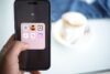 A person holds a smartphone displaying an "AI" folder with various app icons. In the blurred background, a cup of coffee sits on a table, creating a casual tone.
