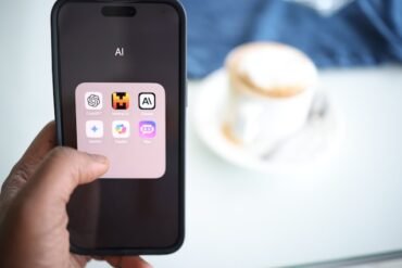 A person holds a smartphone displaying an "AI" folder with various app icons. In the blurred background, a cup of coffee sits on a table, creating a casual tone.