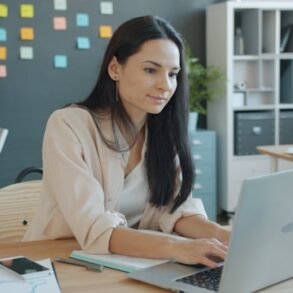 A woman with long dark hair works on a laptop at her desk in a modern office, with a colleague visible in the background.