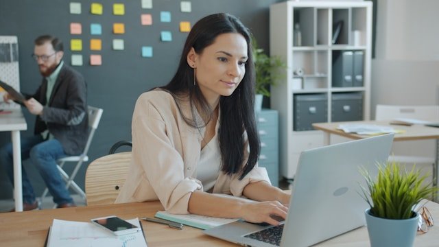 A woman with long dark hair works on a laptop at her desk in a modern office, with a colleague visible in the background.