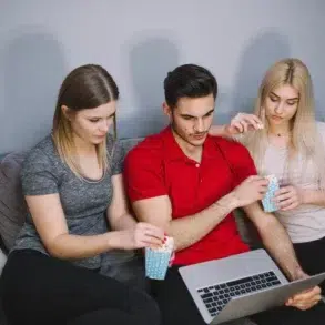 Three friends sit on a couch sharing popcorn while a man in a red shirt uses a laptop.