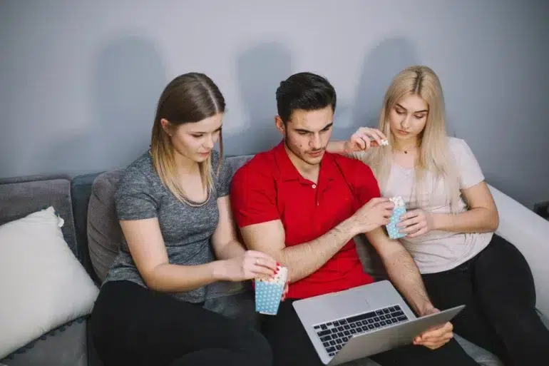 Three friends sit on a couch sharing popcorn while a man in a red shirt uses a laptop.
