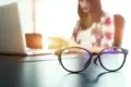 Round eyeglasses sit on a desk beside an open laptop, with a woman reading in the sunlit background.