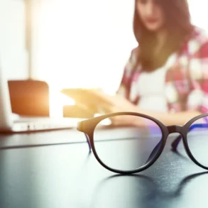 Round eyeglasses sit on a desk beside an open laptop, with a woman reading in the sunlit background.