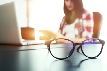 Round eyeglasses sit on a desk beside an open laptop, with a woman reading in the sunlit background.
