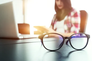 Round eyeglasses sit on a desk beside an open laptop, with a woman reading in the sunlit background.