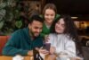 Three friends smile together while looking at a smartphone around a café table with coffee cups and leafy plants in the background.