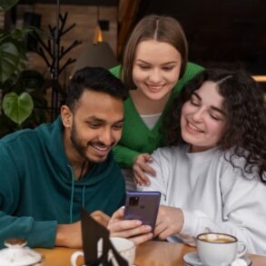 Three friends smile together while looking at a smartphone around a café table with coffee cups and leafy plants in the background.