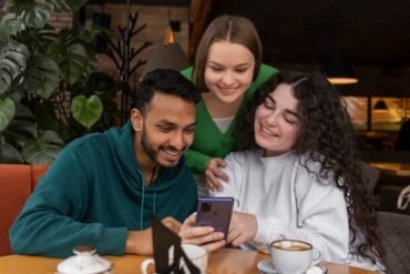 Three friends smile together while looking at a smartphone around a café table with coffee cups and leafy plants in the background.