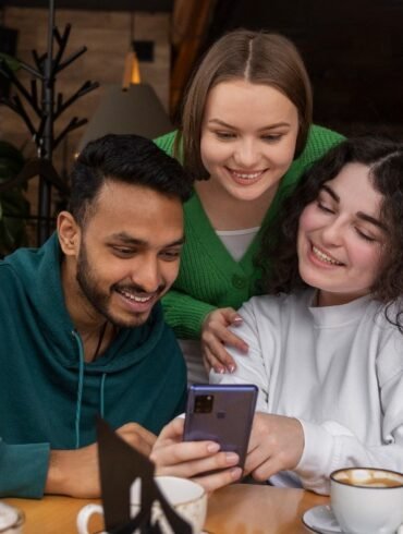 Three friends smile together while looking at a smartphone around a café table with coffee cups and leafy plants in the background.