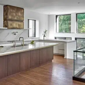 Modern open-plan kitchen with a marble island, wood cabinetry, stainless-steel appliances, and large windows overlooking lush greenery.