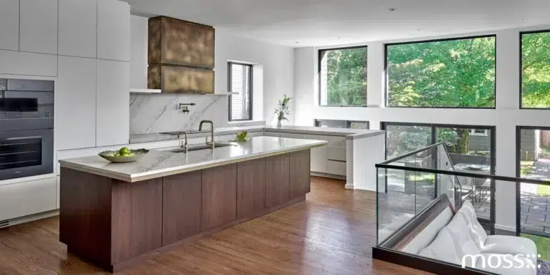 Modern open-plan kitchen with a marble island, wood cabinetry, stainless-steel appliances, and large windows overlooking lush greenery.