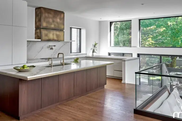 Modern open-plan kitchen with a marble island, wood cabinetry, stainless-steel appliances, and large windows overlooking lush greenery.