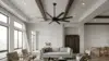 Bright open-plan living room with a gray sofa, armchairs, large windows, a ceiling fan, wood beams, and a dining area.