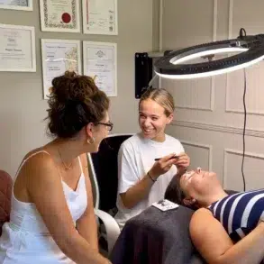 Beautician performing an eyelash treatment on a reclining client under a ring light while another woman observes in a salon displaying framed certificates.