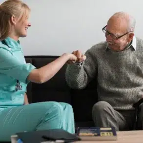 A smiling healthcare worker in teal scrubs fist-bumps an elderly man wearing glasses and a gray sweater while seated on a couch.