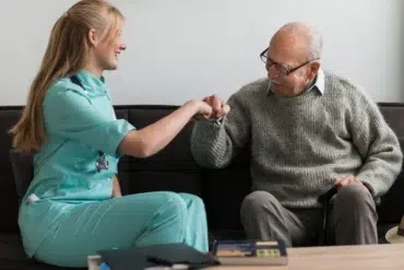 A smiling healthcare worker in teal scrubs fist-bumps an elderly man wearing glasses and a gray sweater while seated on a couch.
