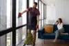 Gloved window cleaner wipes floor-to-ceiling apartment windows while a woman talks on her phone on a blue sofa.