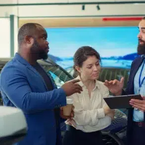 Three adults discuss a car in a showroom, with one man holding a tablet and the woman holding a takeaway cup.