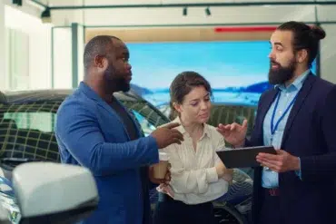 Three adults discuss a car in a showroom, with one man holding a tablet and the woman holding a takeaway cup.