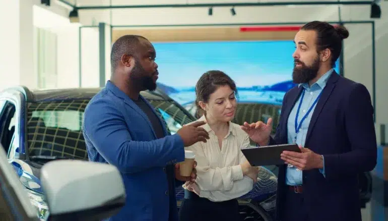 Three adults discuss a car in a showroom, with one man holding a tablet and the woman holding a takeaway cup.