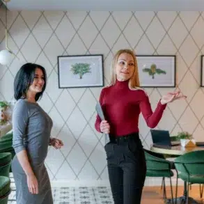 Two women stand in a modern café, with one holding a tablet and gesturing beside tables, green chairs, and framed artwork.
