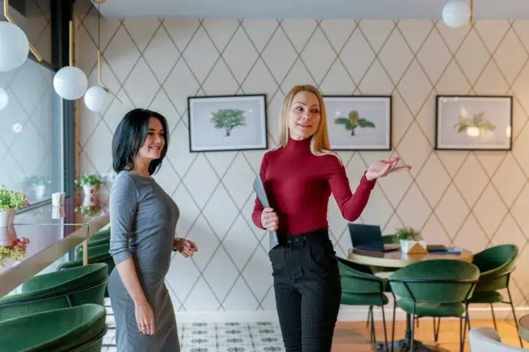 Two women stand in a modern café, with one holding a tablet and gesturing beside tables, green chairs, and framed artwork.