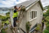 Three workers in high-visibility vests clean leaves from a house gutter, with one standing on a ladder beside the roof.