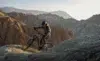 A helmeted cyclist rides a mountain bike along a rocky ridge, with layered, barren mountains stretching across the background.